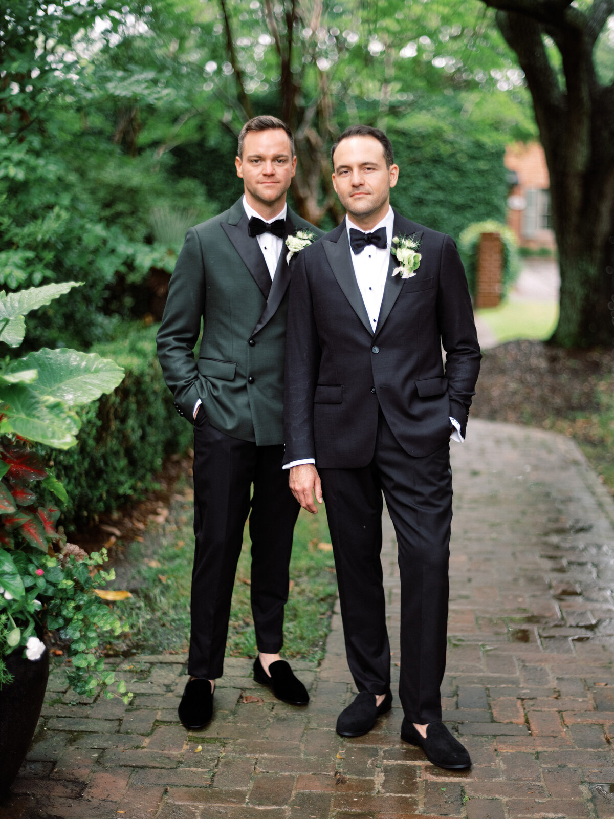 Two elegantly dressed grooms stand confidently in a garden pathway, symbolizing their love for each other amid a romantic and scenic outdoor backdrop of lush greenery.