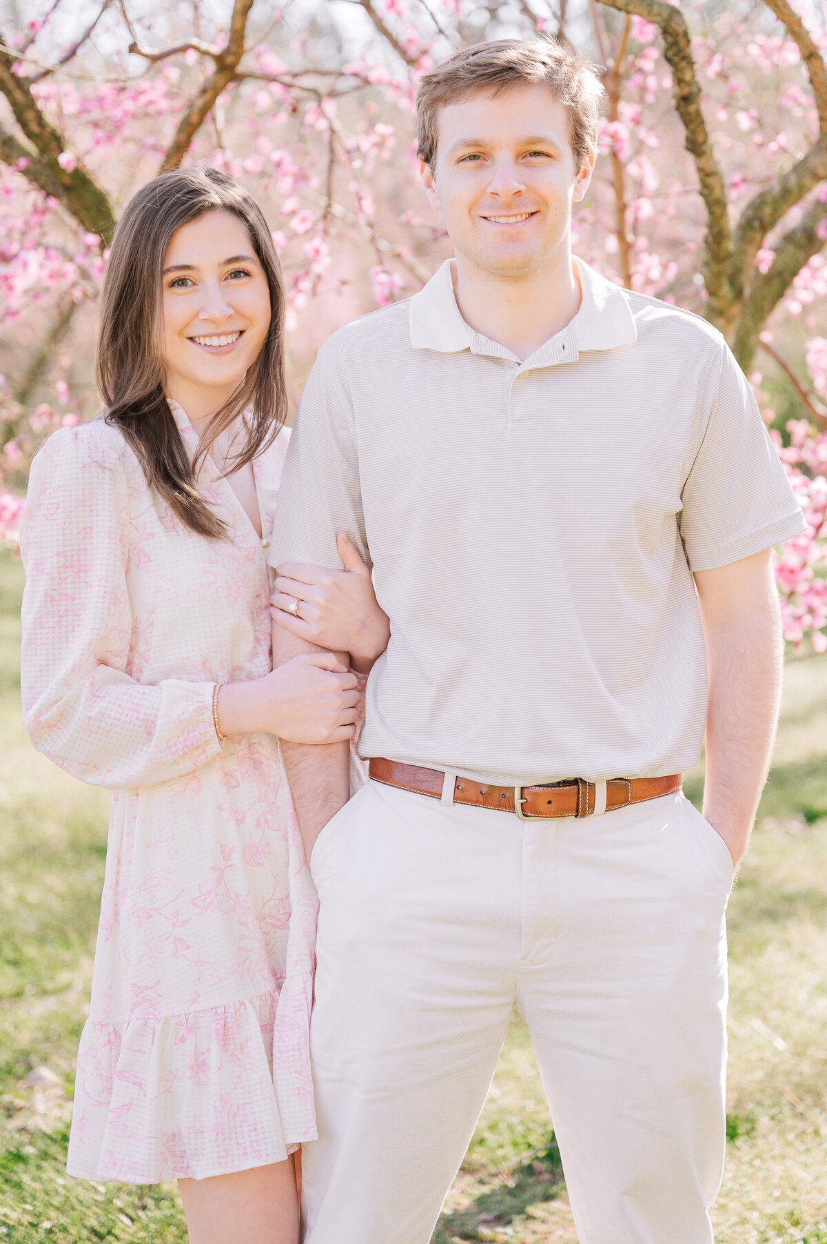 A brunette bride to be holding her soon to be husband along a bright row of pink cherry blossom trees in Raleigh by JoLynn Photography, a Raleigh wedding photographer