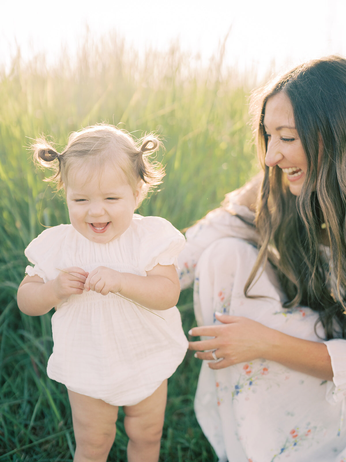 Mother with long brown hair crouches down next to her toddler daughter in pigtails in a grassy field.