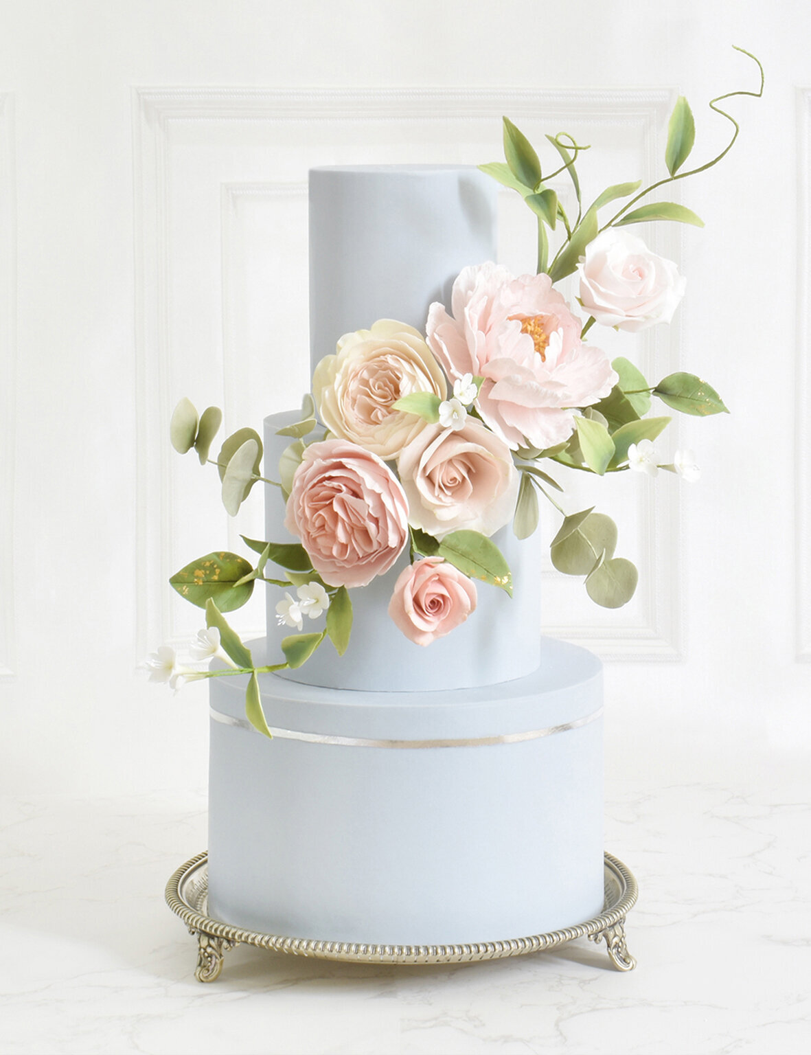a three tiered pale blue wedding cake with a silver leaf band and pink sugar flowers with a panelled wall background