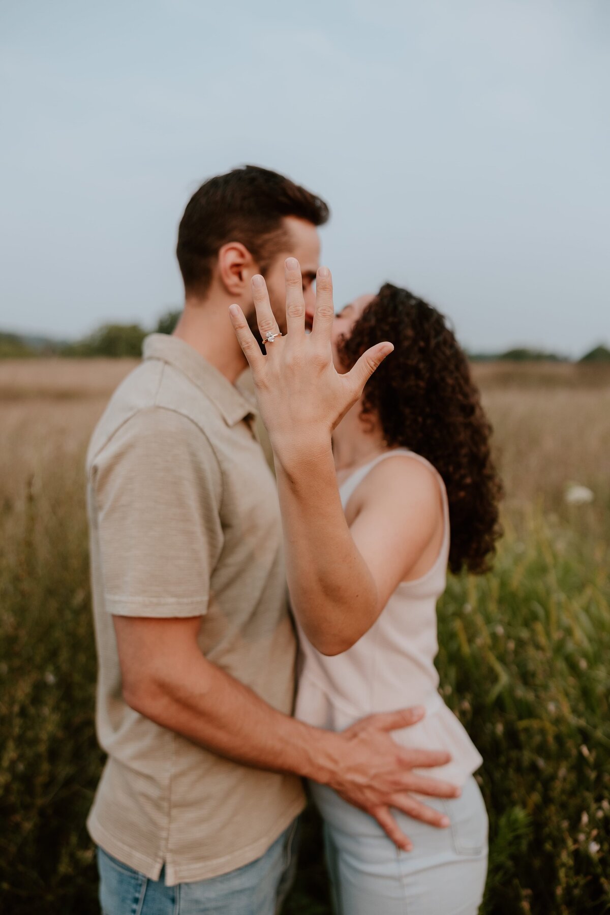 Engaged couple kissing in a field while the bride-to-be holds out her hand to show off her engagement ring in the foreground.