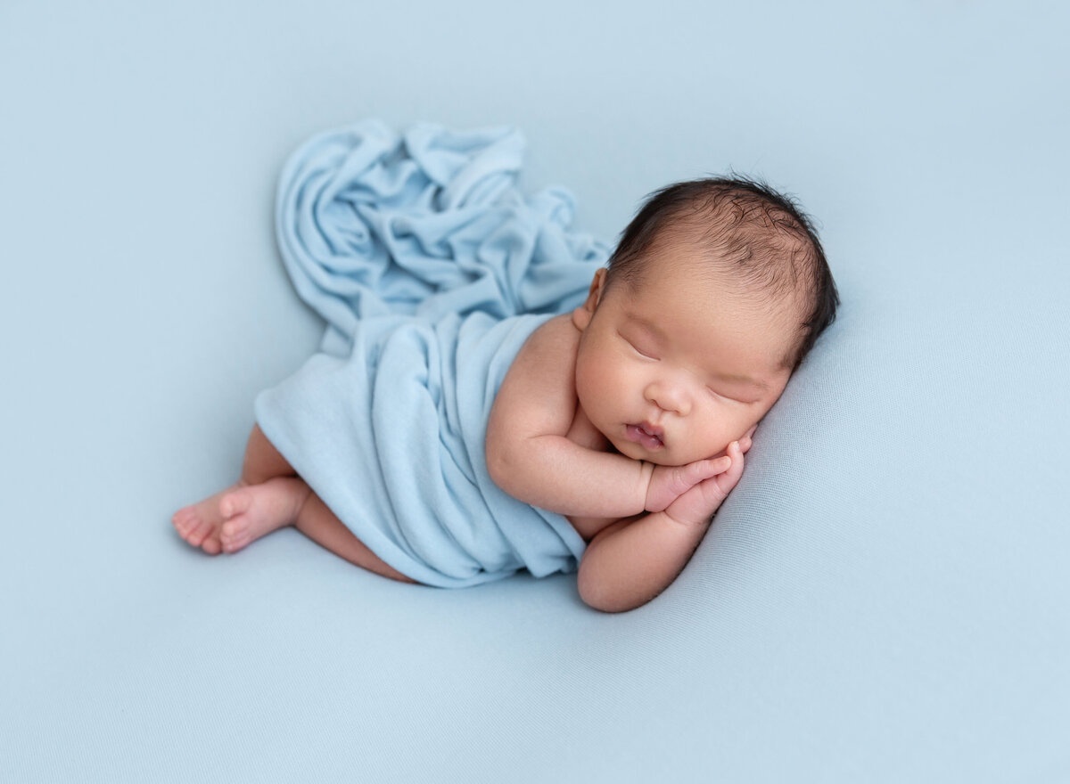 Baby boy sleeping on his side with his hands folded under his cheek. Baby is draped in a blue swaddle fabric.