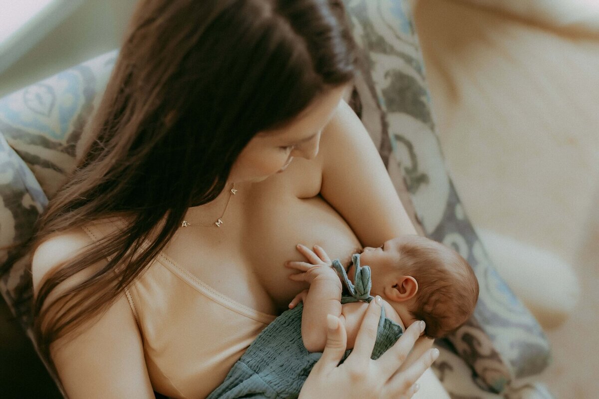 A woman with long brown hair is breastfeeding an infant wrapped in a green blanket, captured in the tender style of a Macon newborn photographer. She sits comfortably on a patterned couch.