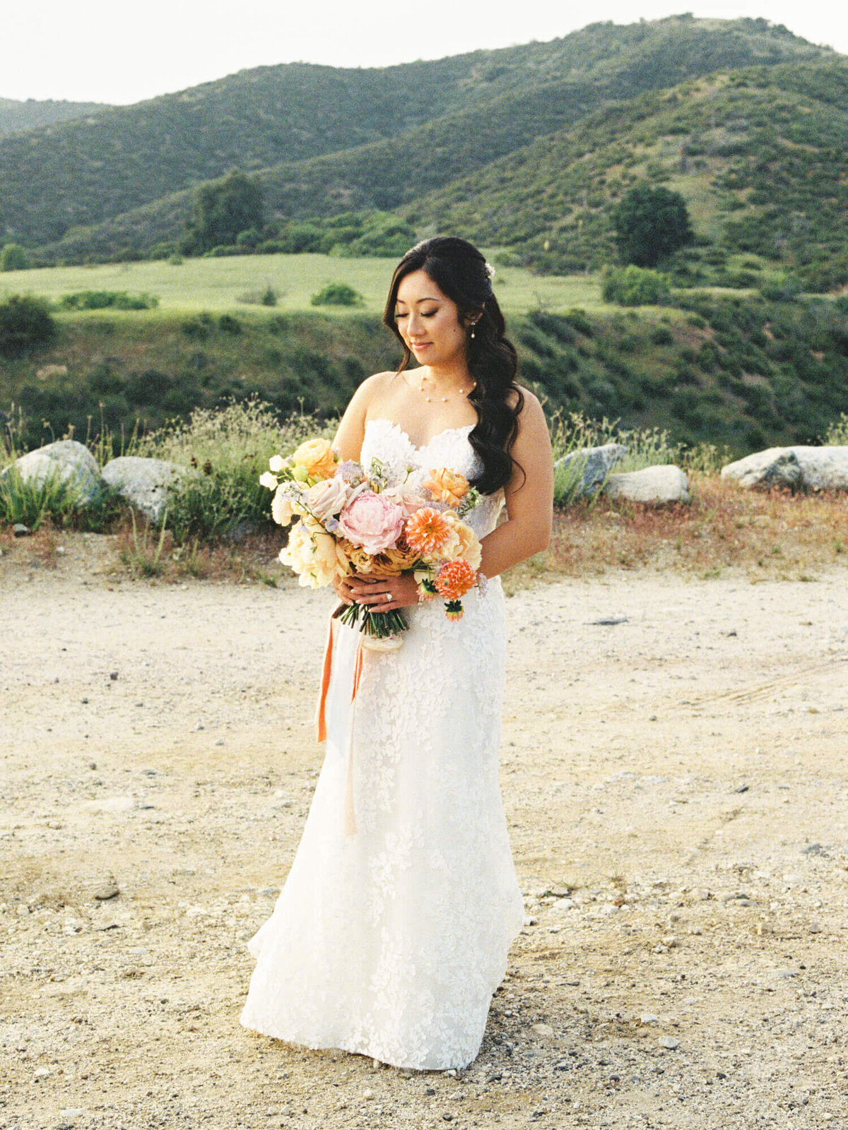 A bride in a lace wedding gown holds a vibrant bouquet, standing on a dirt path with green hills in the background.