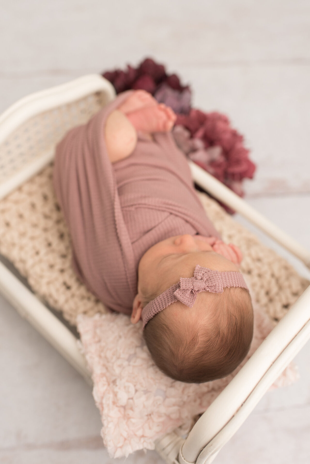 A picture of a newborn girl from the top, lying in a white bed, wrapped in mauve with hydrangeas around her.