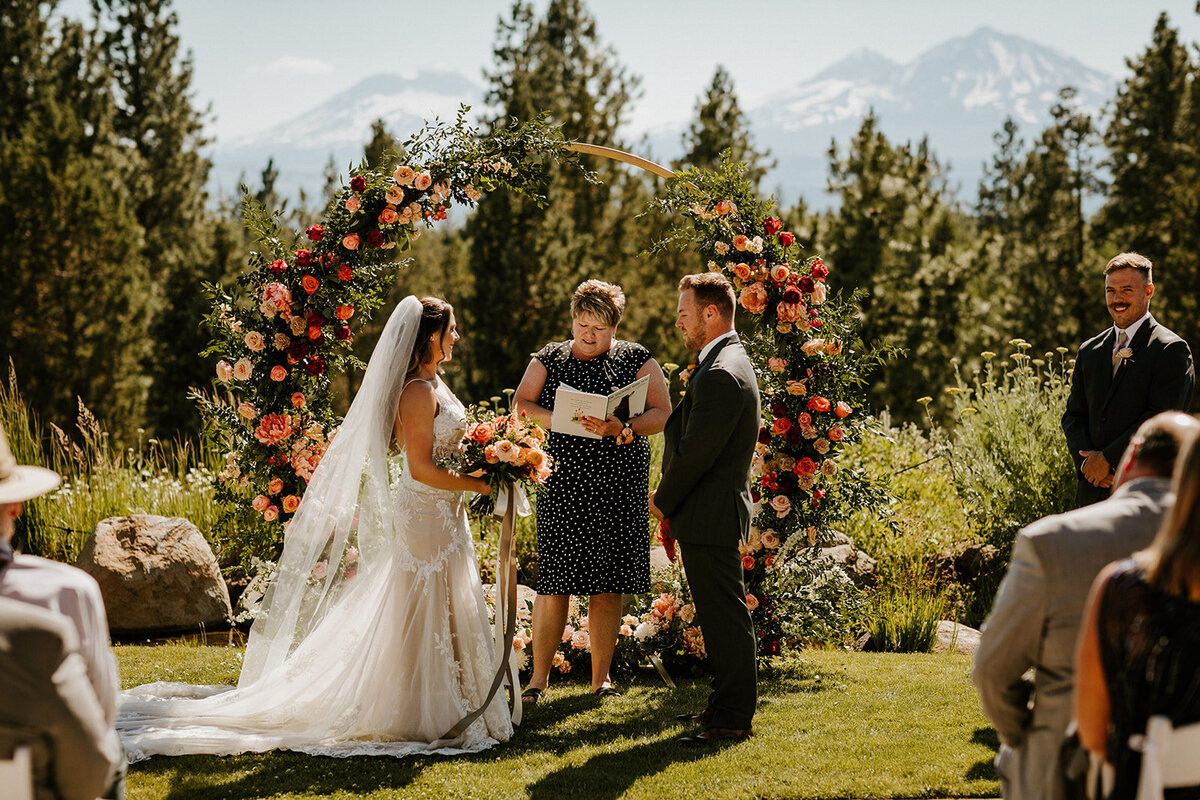 Wedding ceremony in front of coral flowers on gold circle arch with mountain views at Aspen Lakes golf course in Sisters Oregon