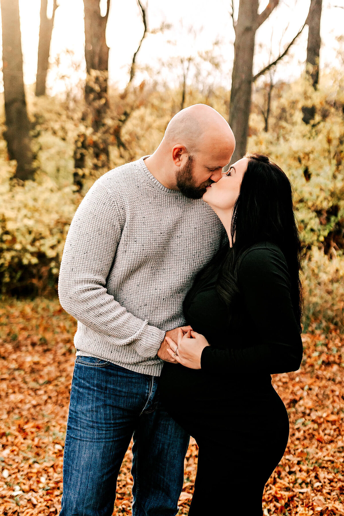 A couple stands outdoors in an autumn forest, holding hands and sharing a kiss. Perfect for maternity portraits, the woman, in a black dress, rests her hand on her pregnant belly as fallen leaves cover the ground.