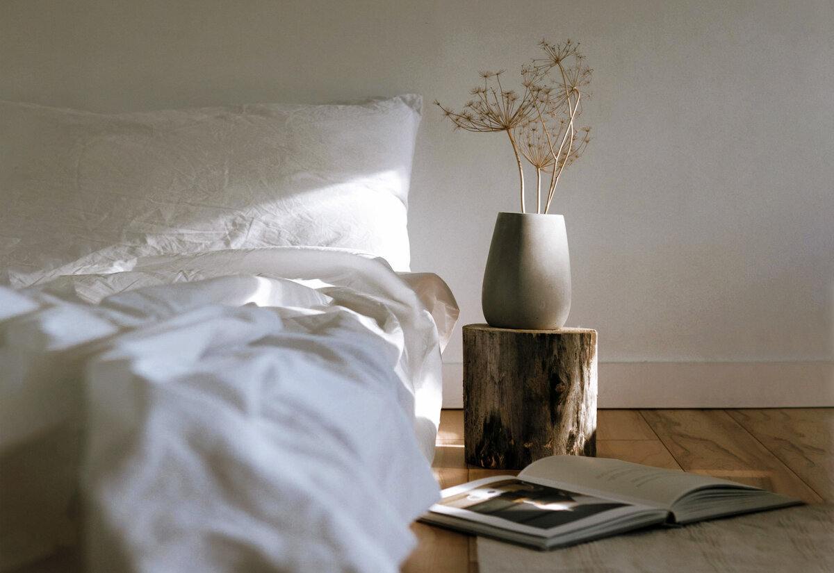 Chambre avec lit de draps blancs, table de chevet en rondin de bois surmontée d'un vase en céramique. Livre ouvert au sol sur un tapis.