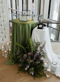 A close-up of a Welcome Area at a National Arts Centre wedding, featuring a green satin linen on a cocktail table with a minimalist black and glass card box and lavender tapered candles. Stacked next to the card box table is a white satin riser with a vintage black telephone used as an audio guest book. A large lavender floral arrangement is placed in front of the tables on the ground surrounded by large floating candles.