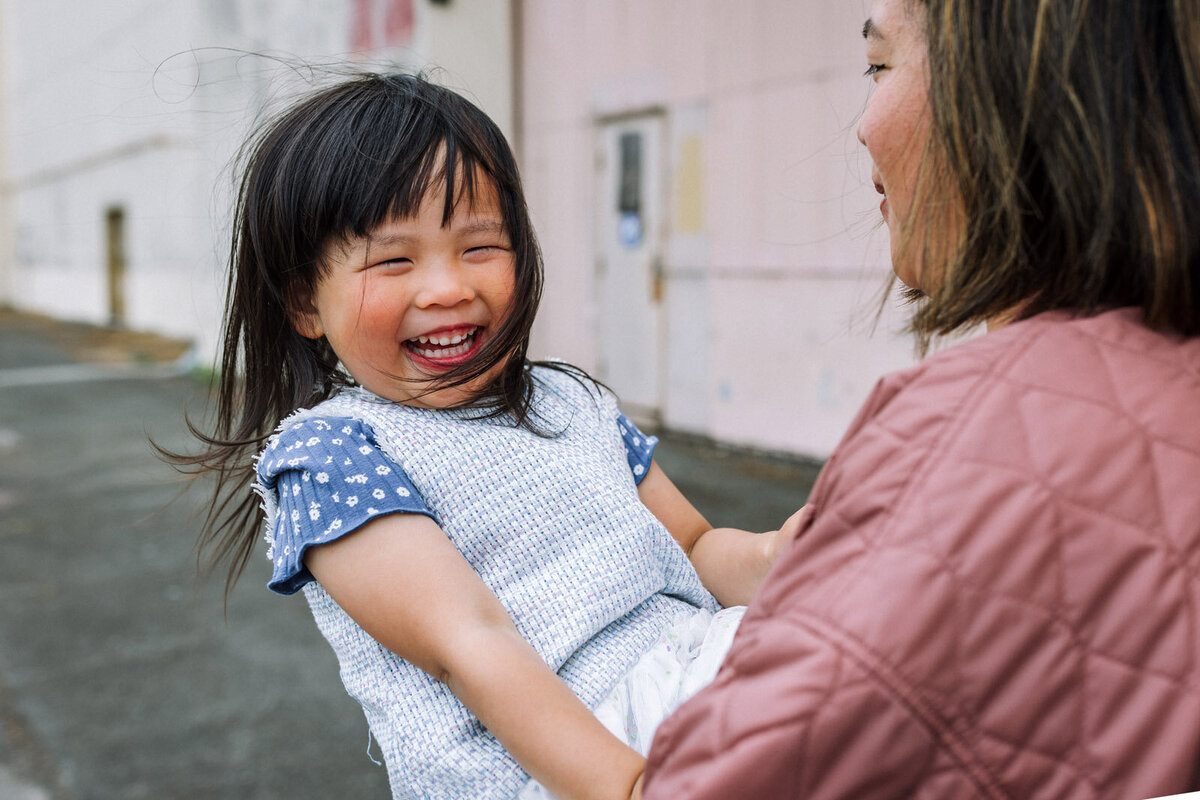 girl laughing in mom's arms as she's spun around and looking  at the camera at Alameda naval base