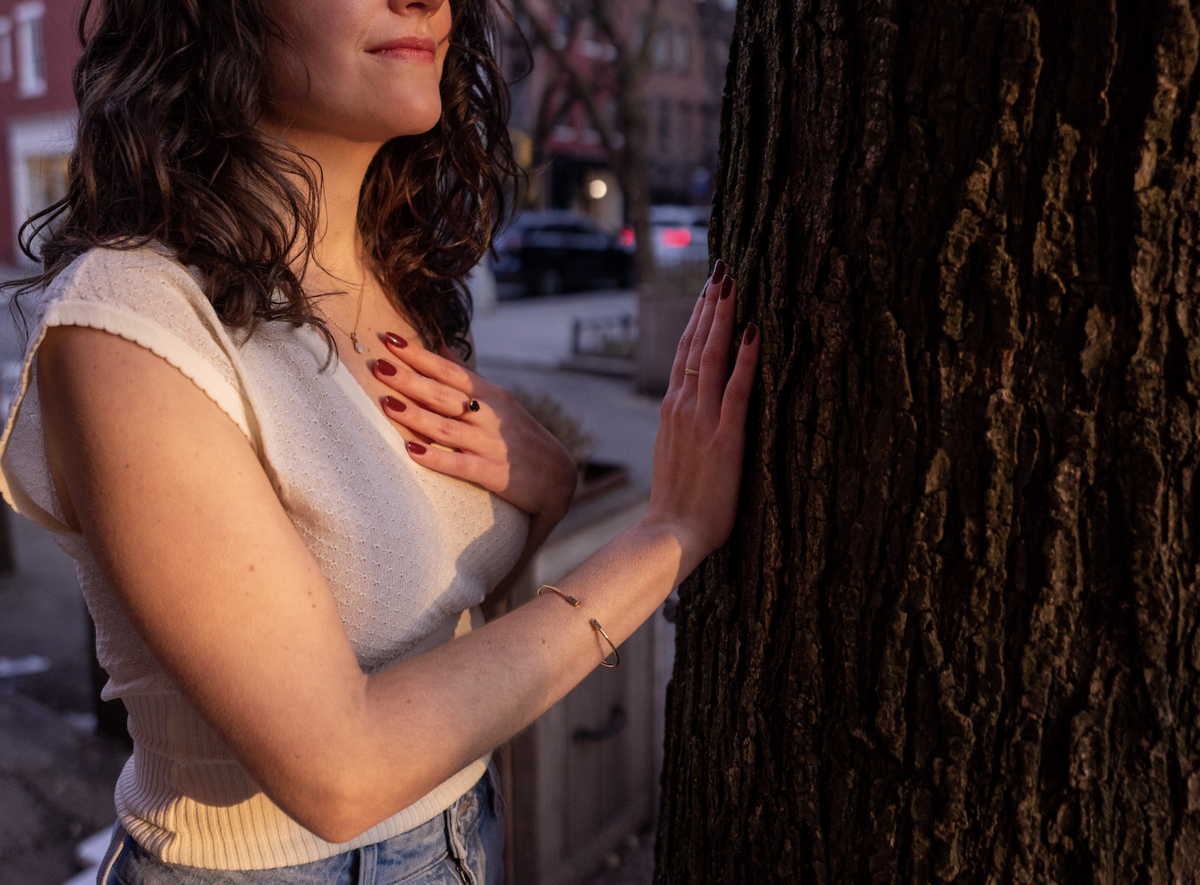 Woman with brown hair smiling and holding one hand on her heart with the other touching a tree