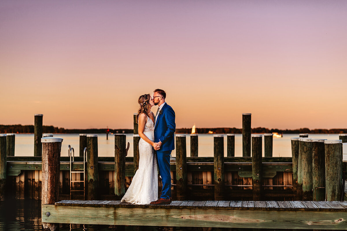 Nautical wedding with bride and groom holding hands and standing on a dock while kissing one another at sunset photographed by Maryland wedding photographer