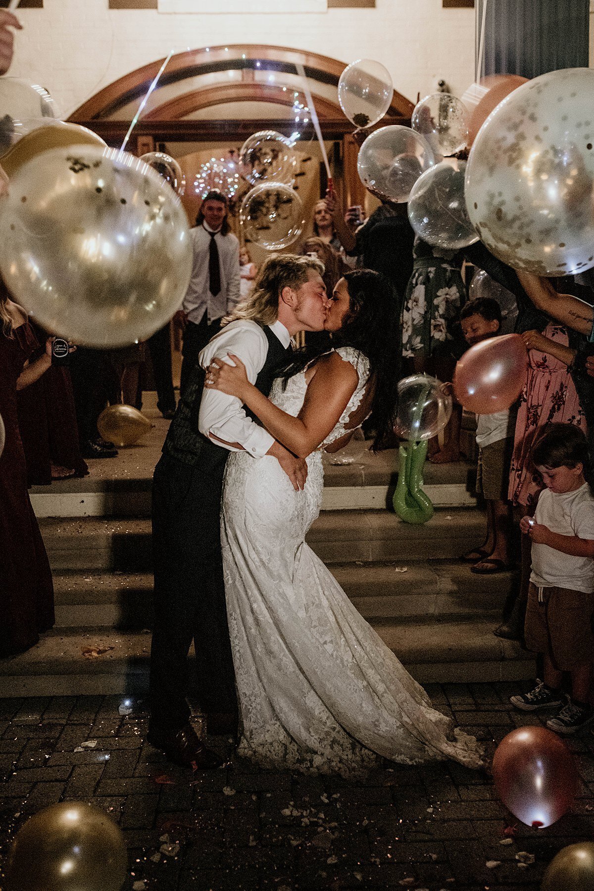 A groom wearing black pants, a black vest and a white shirt dips the bride, wearing a sleeveless wedding dress in a kiss. Wedding guests cheer and shake glitter filled balloons  as they leave their wedding reception.