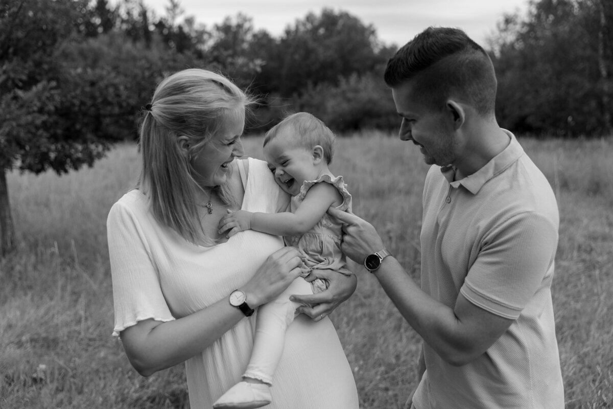 Schwarz weiß Bild von einer Familie, Mutter hat Kind auf dem Arm und der Vater steht daneben, alle lachen