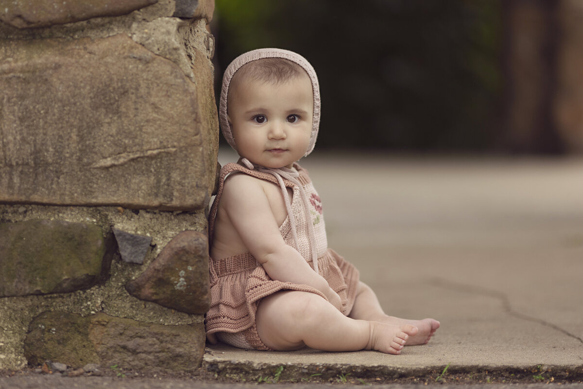 A young toddler girl in a pink dress and matching bonnet sits on a stone step