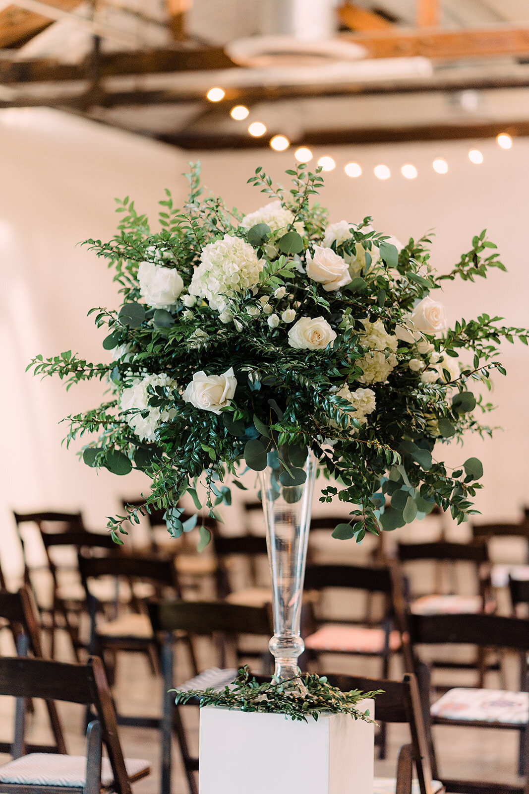 A lush floral arrangement of white roses, hydrangeas, and green foliage set in a tall glass vase, ready to adorn the ceremony space with chairs arranged in rows.