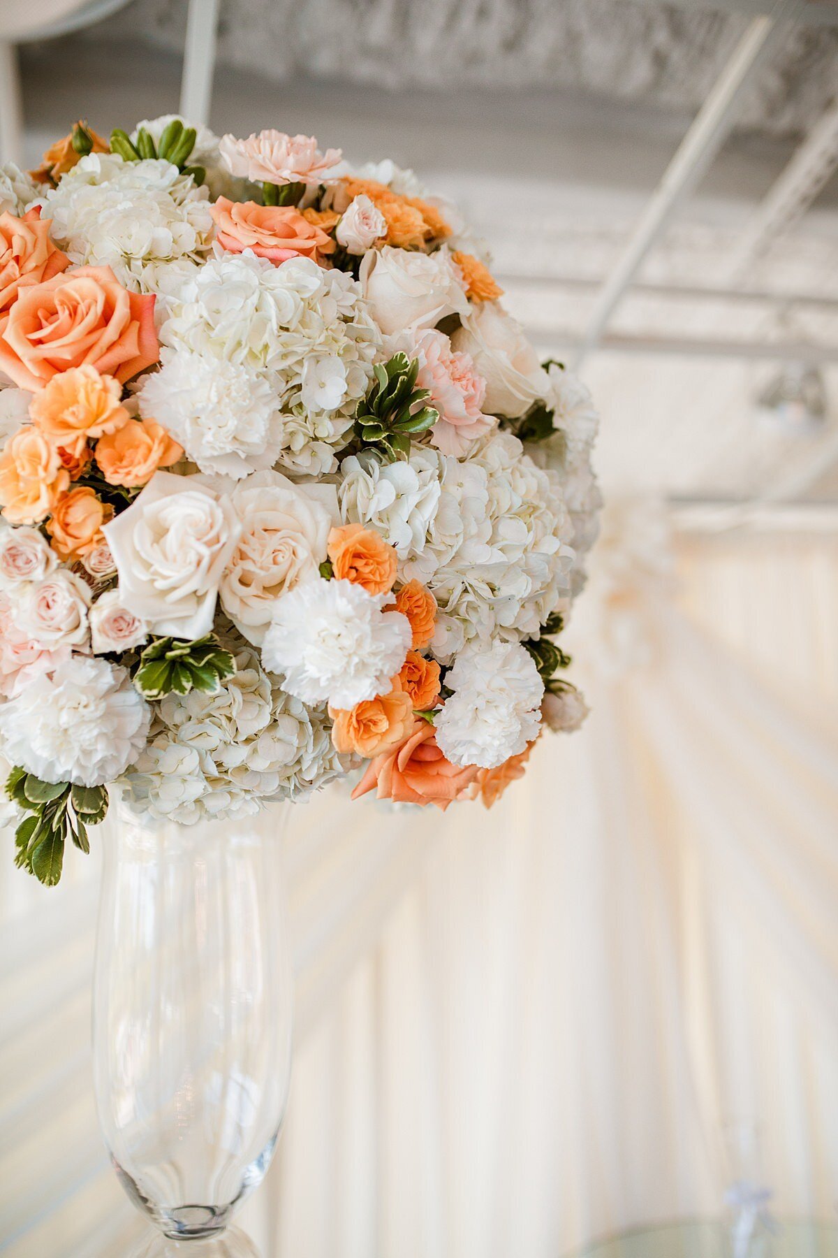 Sitting on top of a tall glass vase in front of a white draped wall at The Liff Center is a large round floral centerpiece. the centerpiece consists of white hydrangea, ivory roses, white carnations, peach tea roses, orange roses, blush tea roses and hints of white tipped greenery.