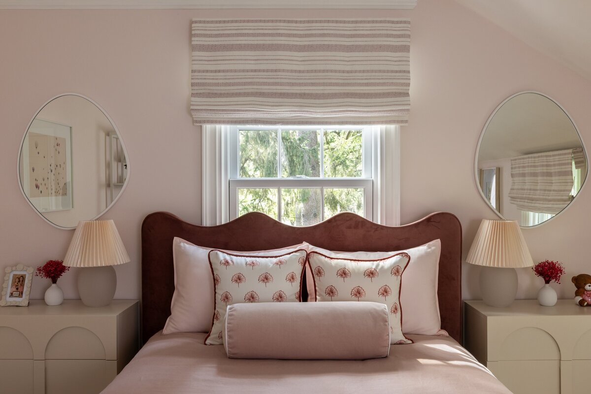 Cozy and elegant girl's bedroom featuring a wave-edged velvet headboard, blush pink bedding, scallop-printed pillows, and a soft roman shade over a double window. Matching oval mirrors and fluted lamps sit on modern cream nightstands, adding symmetry and charm to the space.