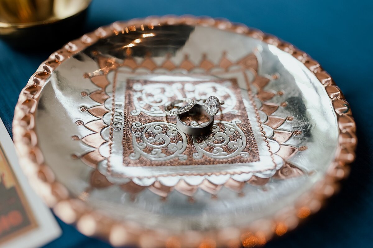 Oval cut diamond engagement ring, diamond infinity band and platinum wedding band sitting on a silver and copper hand hammered plate for an Indian wedding in Nashville, TN