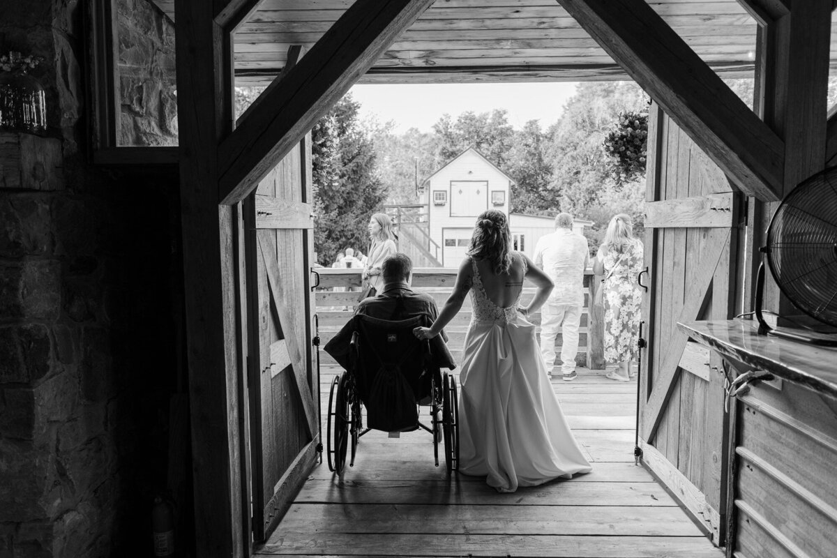 Black and white photo of a bride in the doorway of a rustic barn with a guest in a wheelchair in the foreground