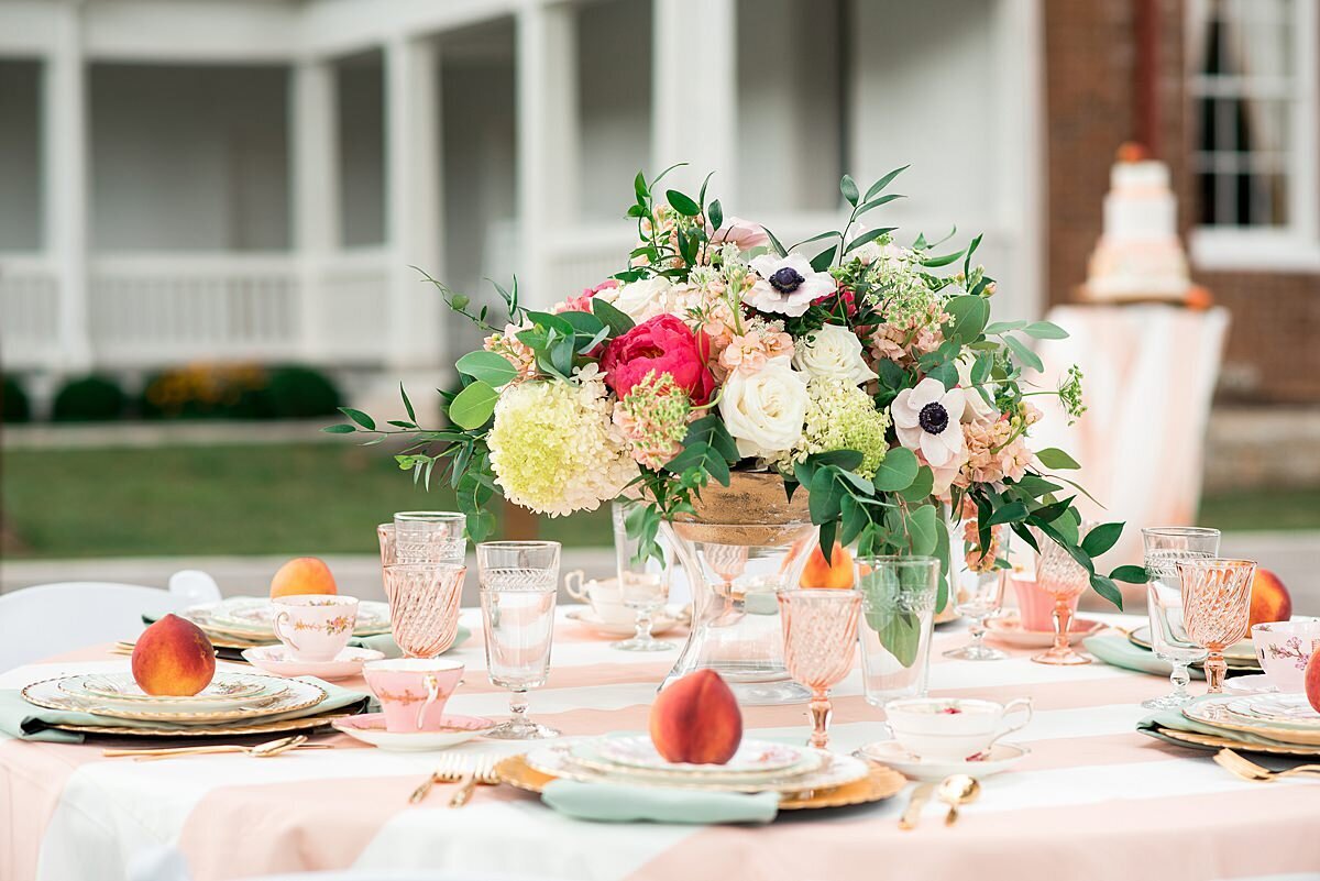 Large floral centerpiece in a gold footed bowl with yellow, ivory, white, peach  and pink flowers on a peach and white striped table cloth. at each place setting on top of a gold fluted charger with a sage green linen napkin is a set of three mismatched vintage china plates topped with a fresh peach and accepted with pink footed vintage water goblets.
