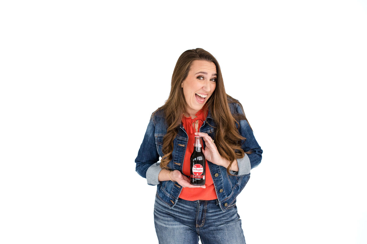 brand photo for a soda brand with woman in a denim jacket and pants posing with a bottle on a white background
