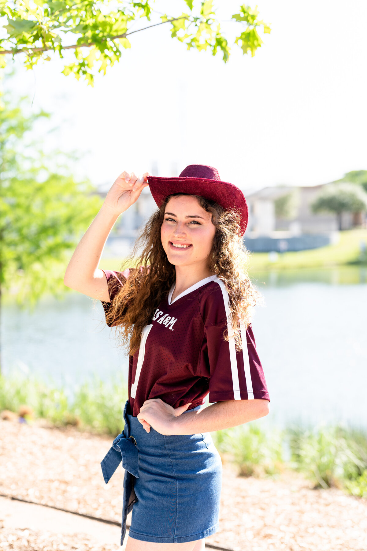 Texas A&M senior girl with hand on hip and maroon cowboy hat smiling while wearing Aggie maroon jersey and jean skirt at Aggie Park
