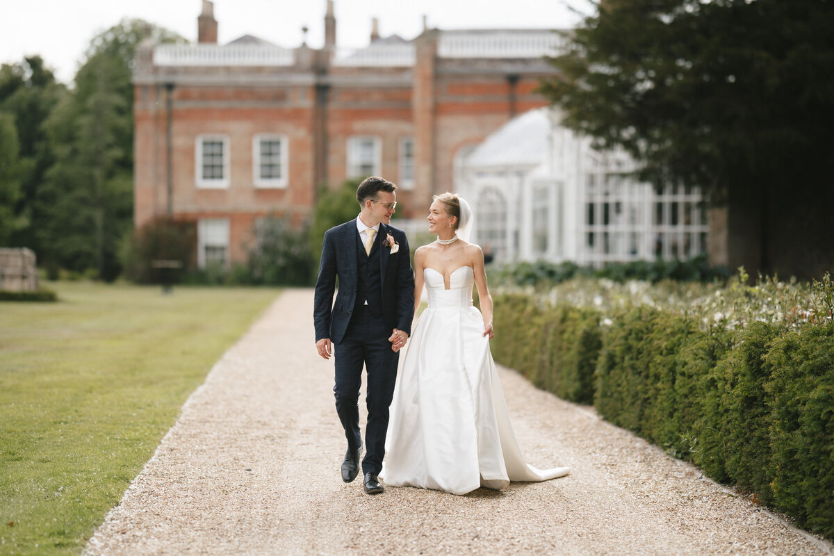 A bride and groom walk hand in hand down a gravel path, smiling at each other during their Avington Park Wedding. The bride wears a white strapless gown; the groom is in a dark suit. A large brick building and garden are in the background.