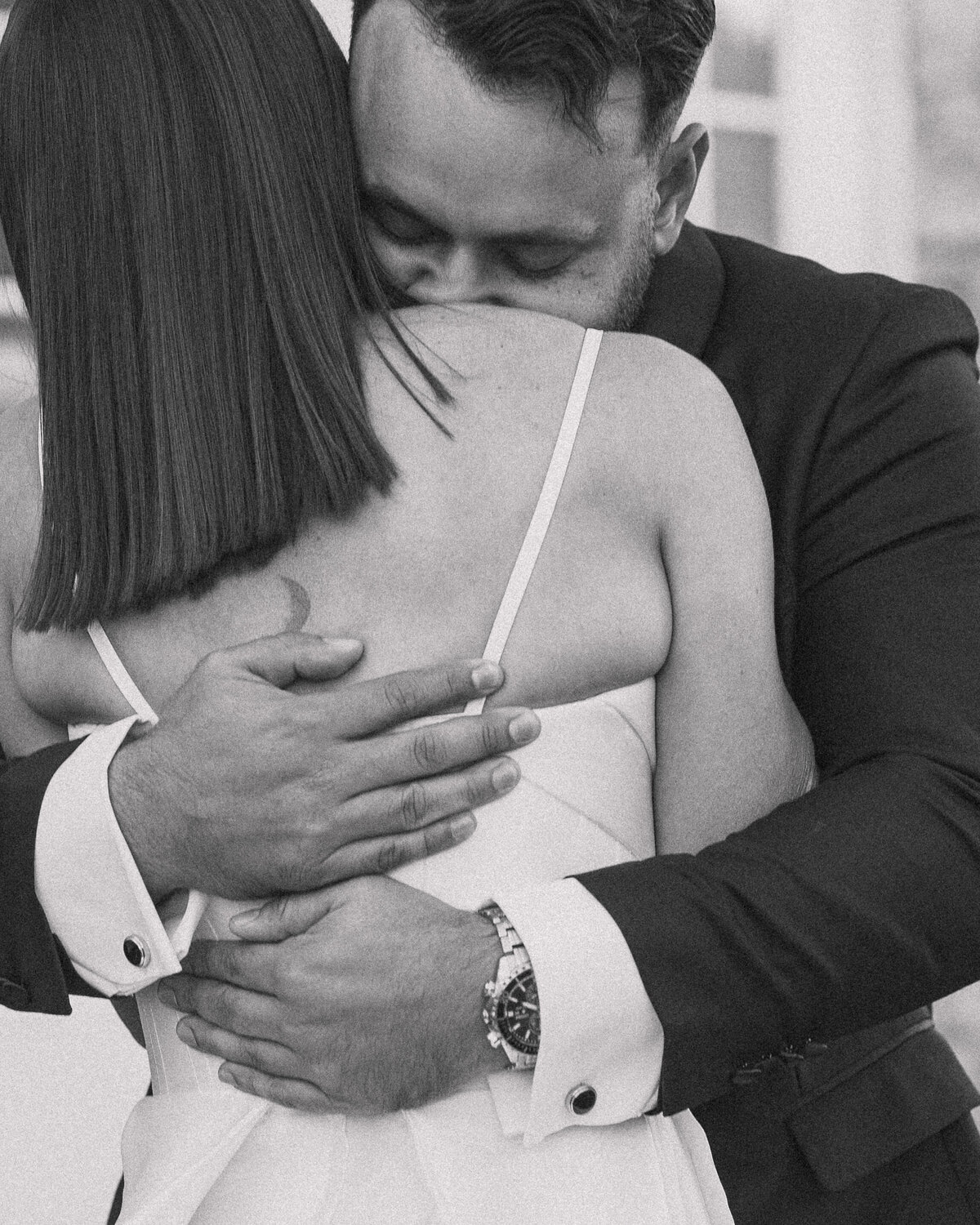Intimate black and white close-up of a wedding couple embracing. The groom, dressed in a suit, tenderly hugs the bride, who wears a sleeveless dress. The groom's face is nestled against the bride's shoulder, conveying deep emotion and affection. His hands, adorned with a watch and cufflinks, rest gently on her back, capturing a moment of closeness and love.