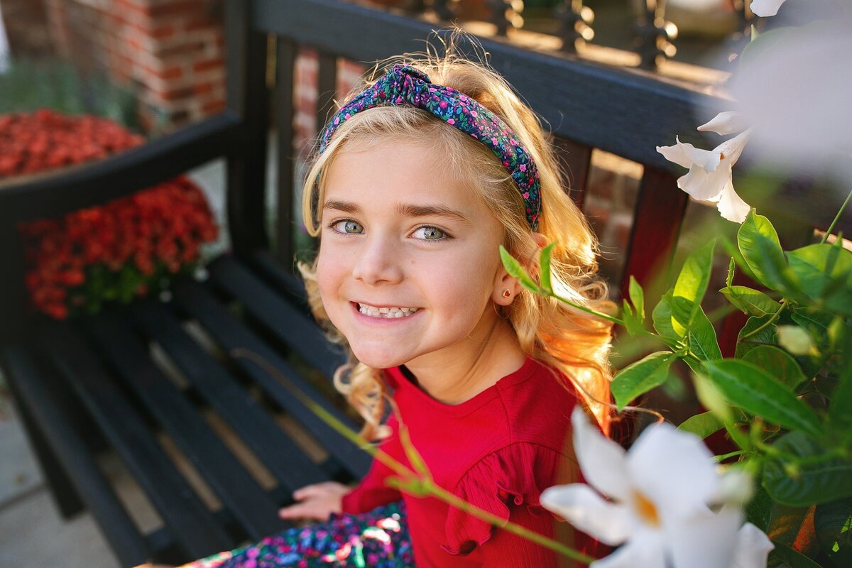 A-young-girl-with-blonde-curls-and-a-colorful-headband-smiling-on-a-black-bench-with-flowers-in-downtown-Zionsville-Indiana-during-a-fall-family-photo-session-capturing-the-small-town-charm