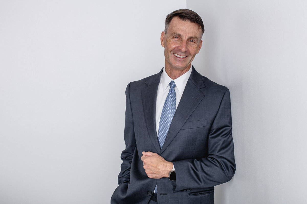 A man with brown hair wearing a gray suit and a blue tie standing against a gray wall with his left arm on his suit jacket