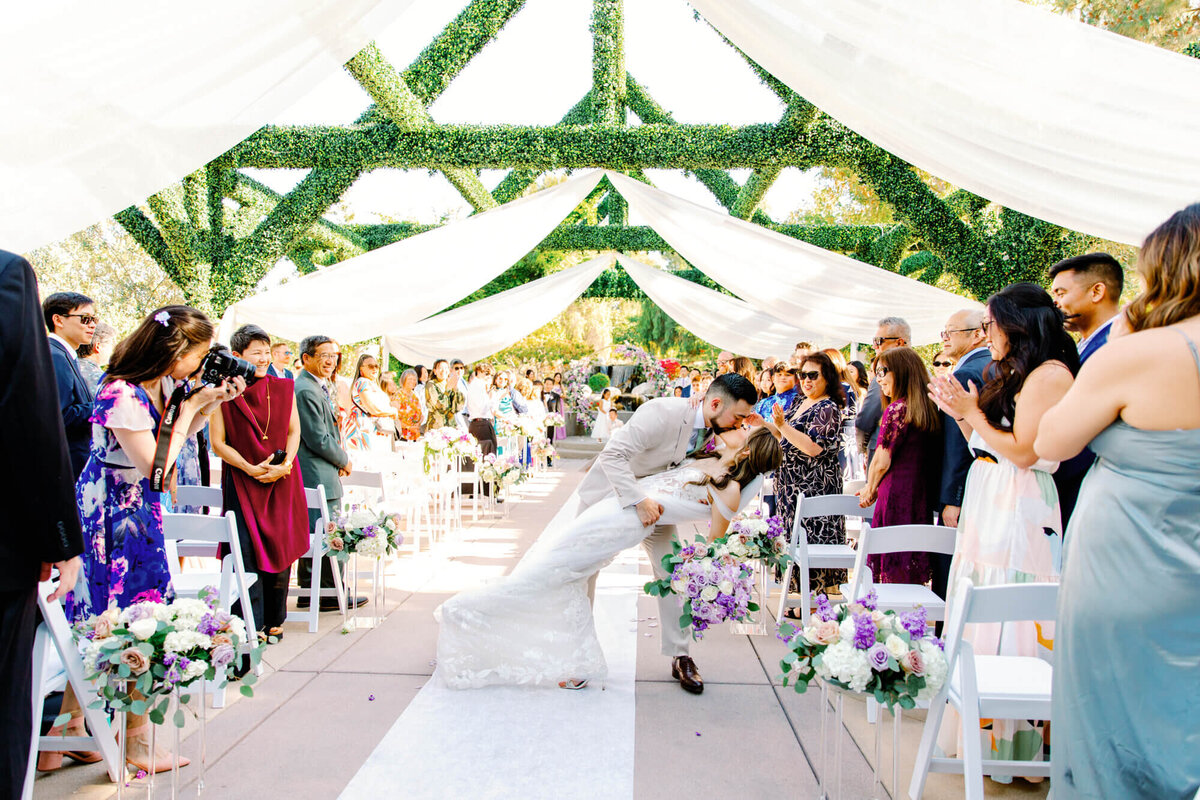 Bride and groom share a romantic kiss under white draped fabric and greenery in a sunlit aisle, surrounded by applauding guests and flowers.