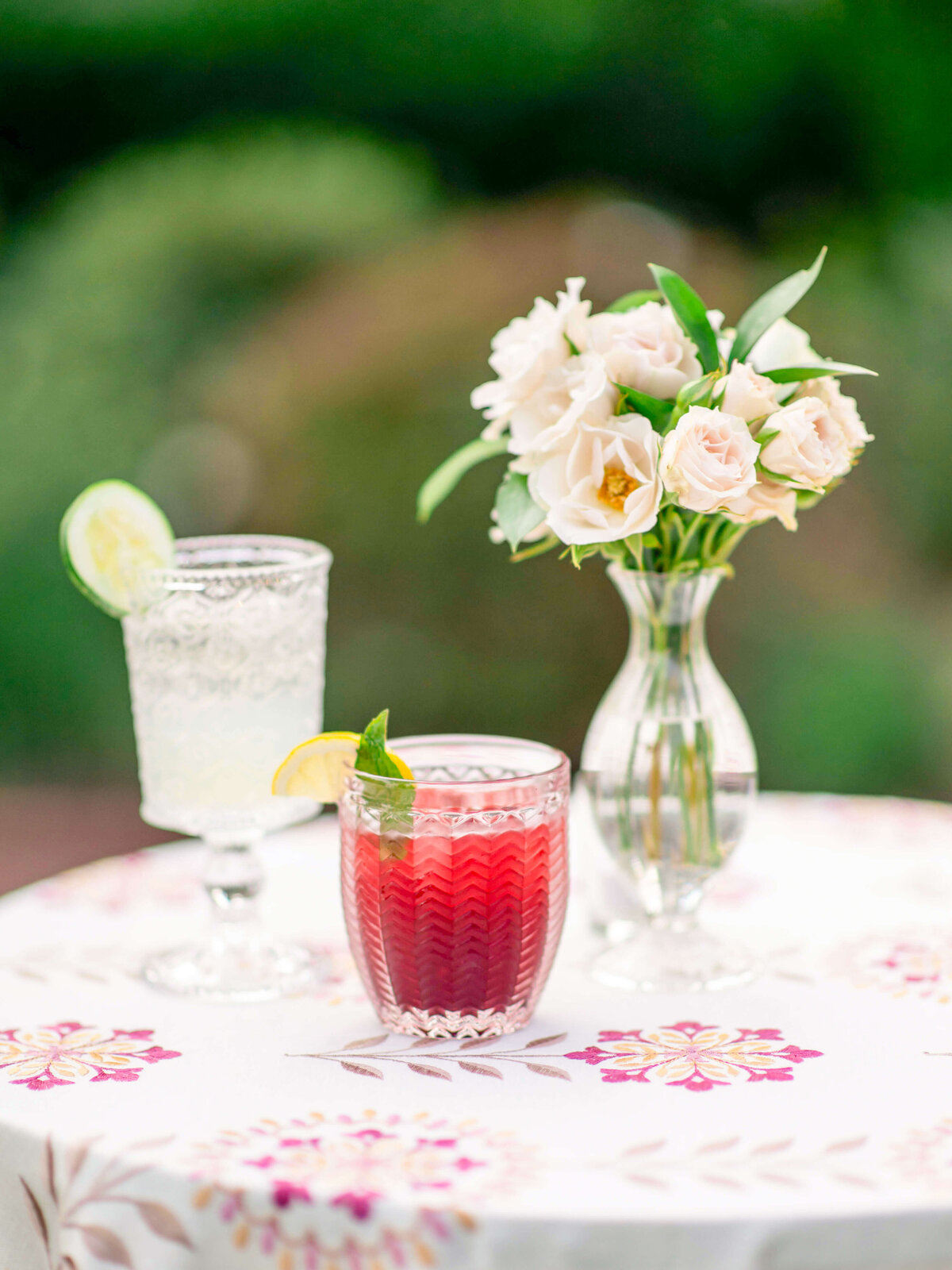 Two glasses with beverages and garnishes on a floral tablecloth: one with a red drink and mint, the other with a clear drink and a lime slice. A small vase with white roses is placed beside them. The background is a blurred greenery.