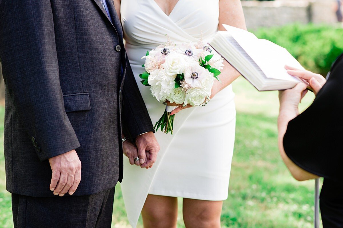 Detail image of a wedding ceremony at Cheekwood. The groom and bride hold hands. The groom is wearing a dark gray suit and the bride is wearing a short white dress with a plunging neckline and an asymmetrical  skirt. She is holding a large bouquet of white flowers with blush accents. The officiant, dressed in black, reads from a book.