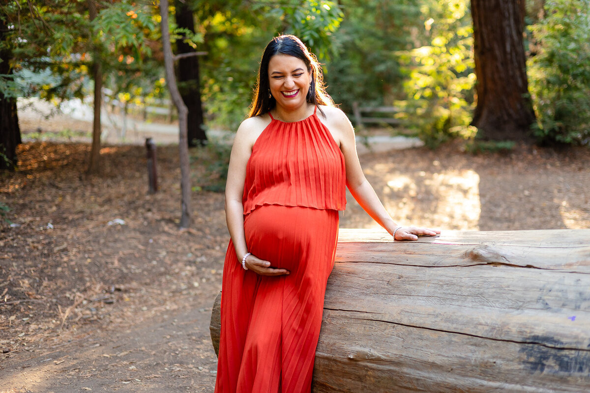 Smiling pregnant woman posing by a large log in a rust-colored pleated dress – Ellobelle Photography