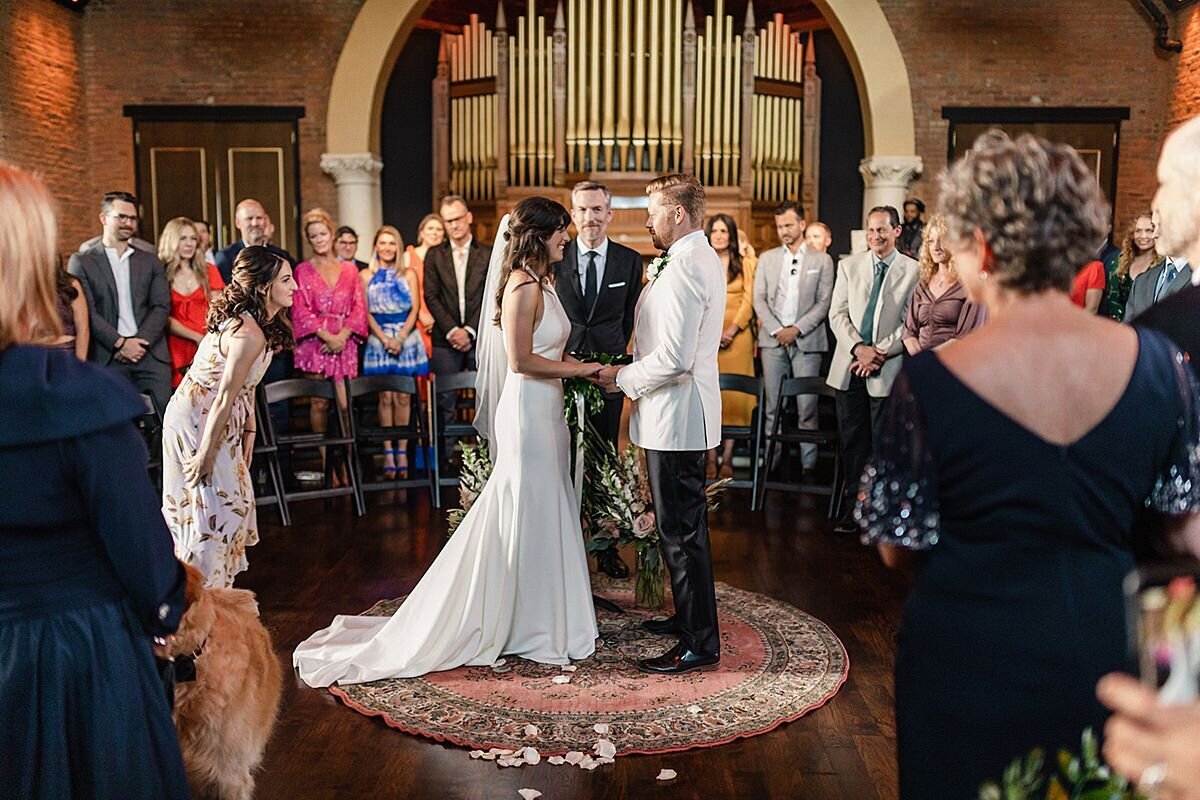 The bride, wearing a silk halter top sheath dress and the groom wearing a tuxedo with a white jacket, exchange vows in the round at their circular set up wedding ceremony in front of the historic pipe organ at Clementine Hall