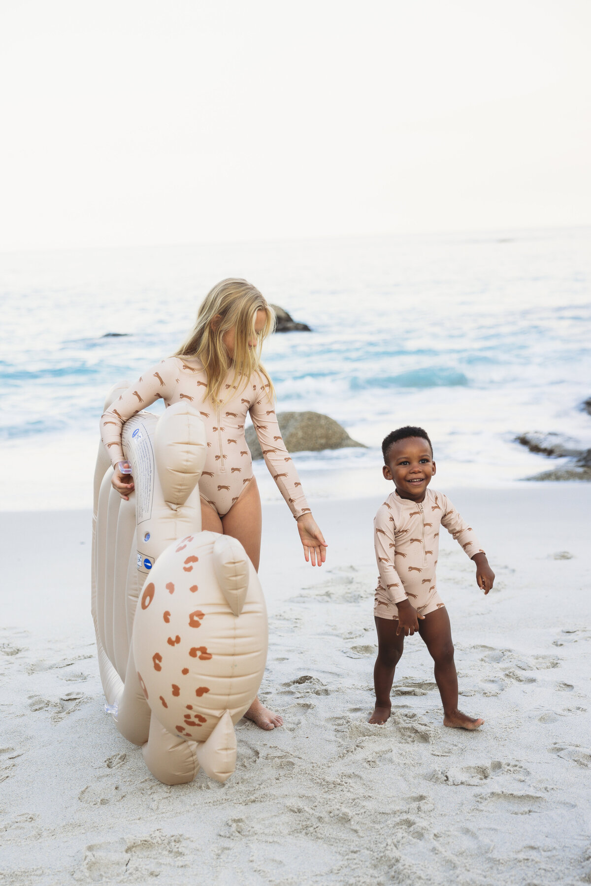 Smiling boy and girl in matching beige swimsuits with playful cheetah prints, enjoying a beach day with inflatable sea animal floats near the shoreline.