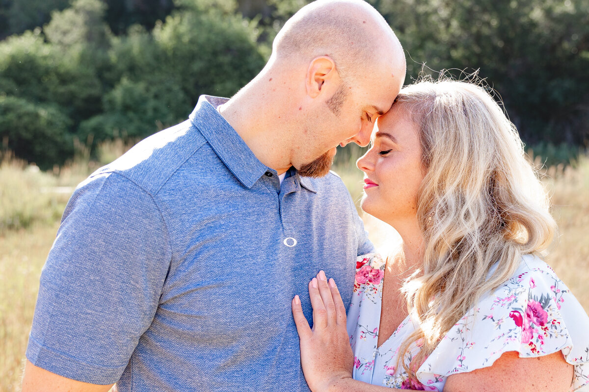 A couple stands closely in a sunlit field, touching foreheads as they embrace. The warm afternoon glow highlights their affectionate connection, beautifully captured by Cassie Lee Photography.