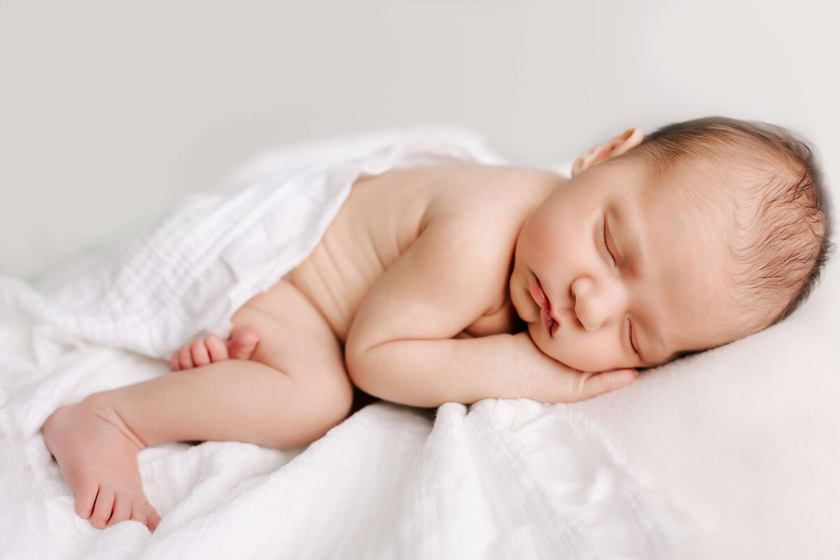 Newborn sleeping on a white blanket while loosely wrapped.