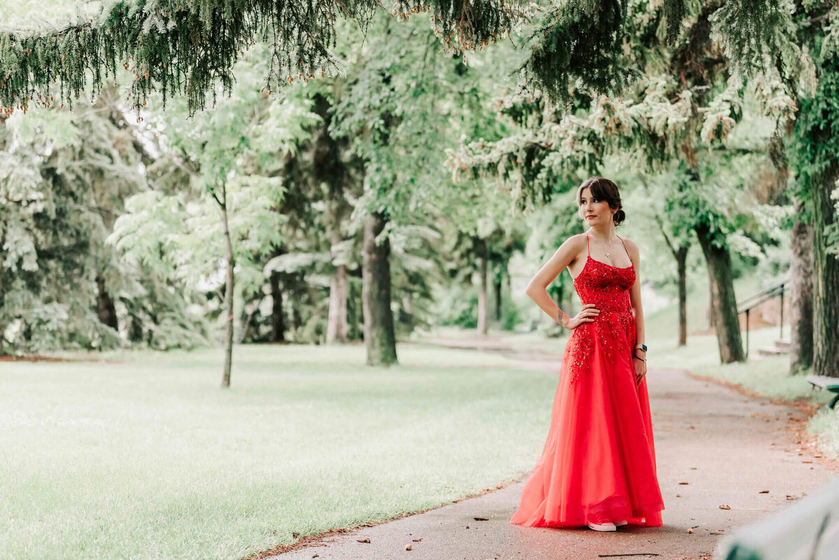 short haired beaty wearing a red dress  is standing on a path with trees nearby. At Saskatoon Saskatchewan.