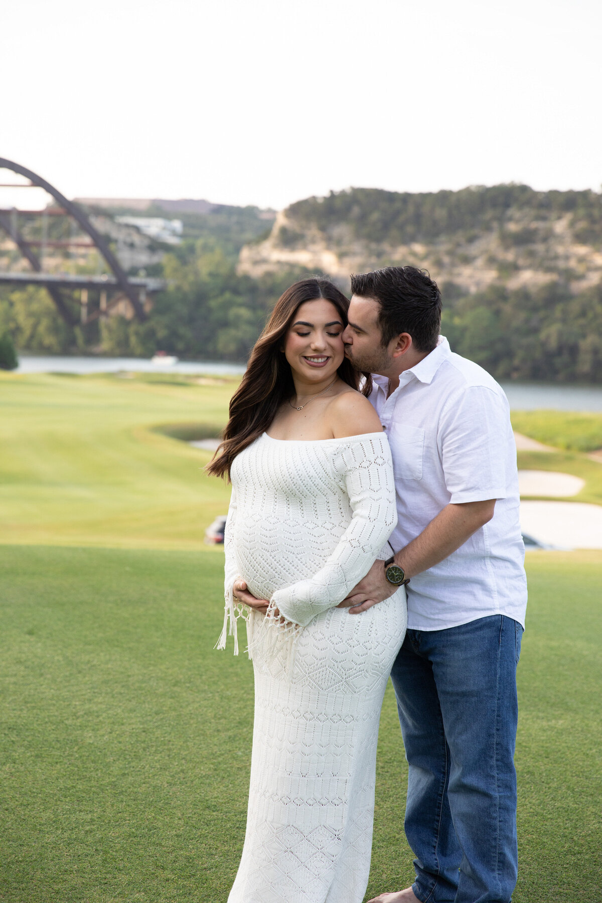 An Austin wedding photographer captures a heartfelt moment as a pregnant couple shares a tender kiss in front of a bridge near a picturesque golf course.