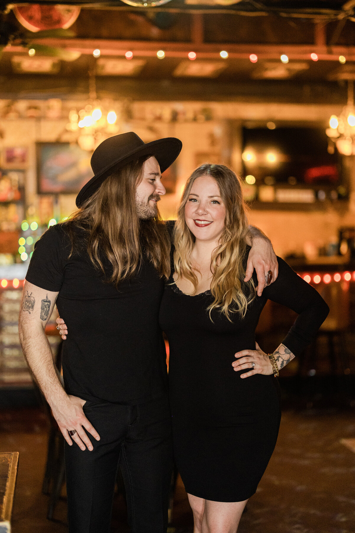 A couple smiling and holding each other close in a bar during their engagement session in Fort Worth, Texas. The woman on the right is wearing a short, black dress while the man on the right is wearing a black shirt, black pants, and a black hat. They both have tattoos on their arms.