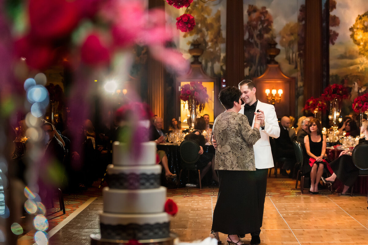 wedding photo peeking through red roses and cake to see mother and son dancing