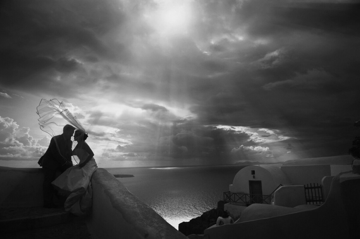 Silhouetted couple kissing with a veil blowing in the wind under dramatic cloudy skies overlooking the ocean. This photo was taken by a destination wedding photographer for Santorini sunset weddings