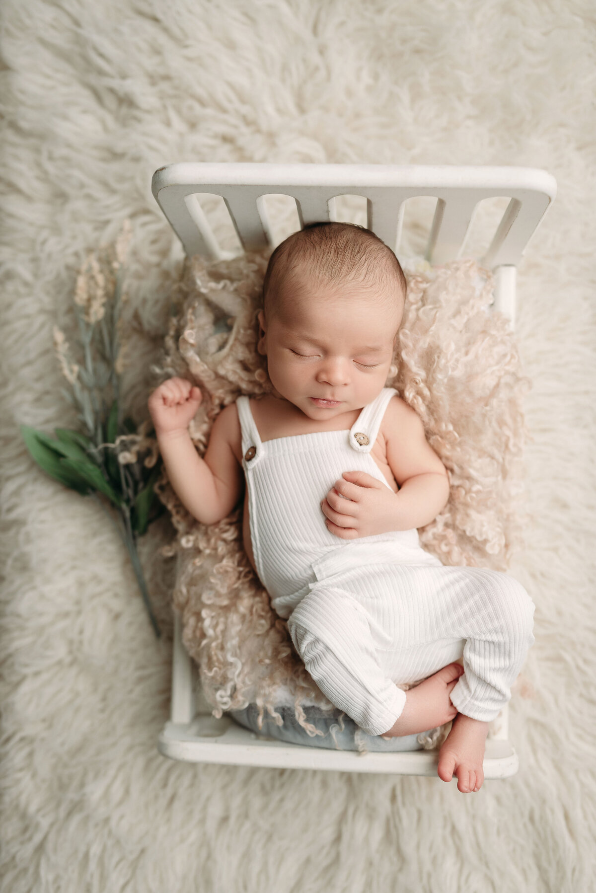 Baby boy wearing white romper laying on cream fur in miniature white baby bed with greenery underneath