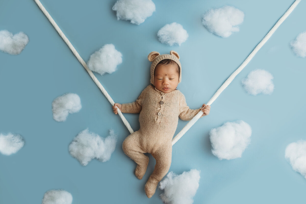 Baby boy in bear outfit holds onto a rope swing amongst the clouds.