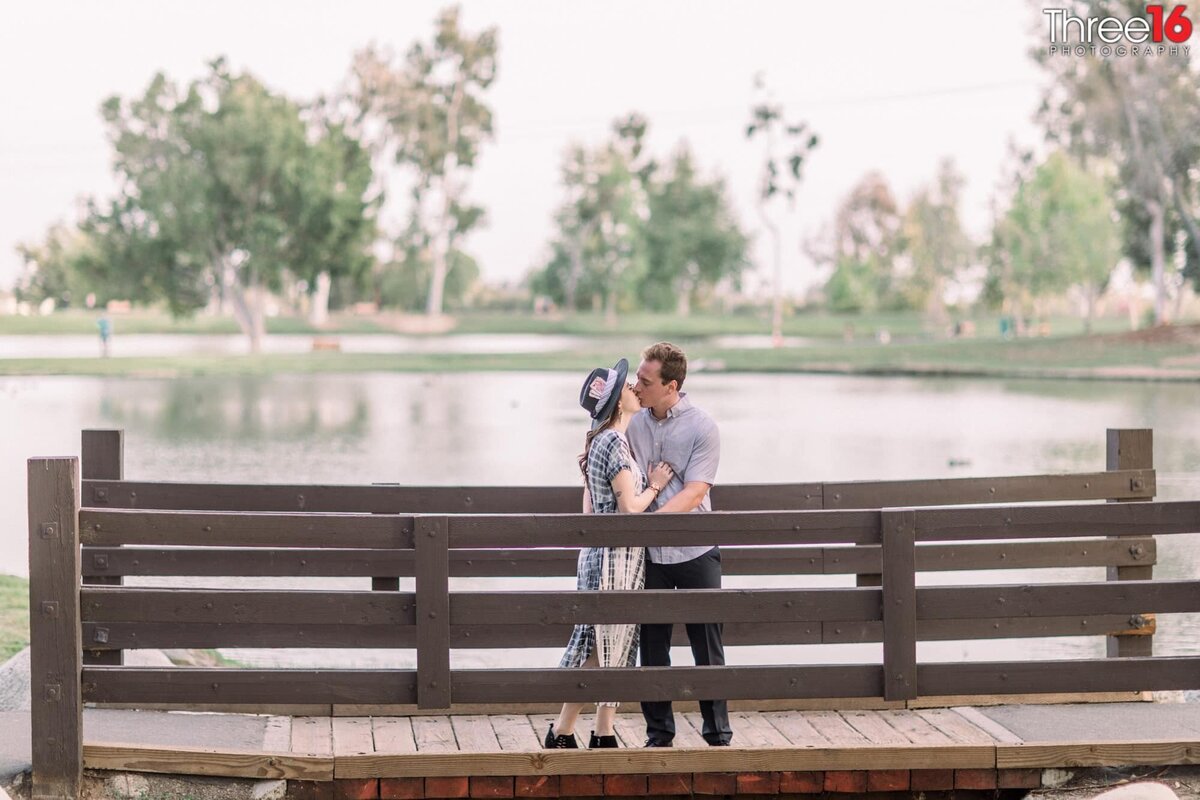 Engaged couple engage in a kiss while on a wooden bridge over the lake
