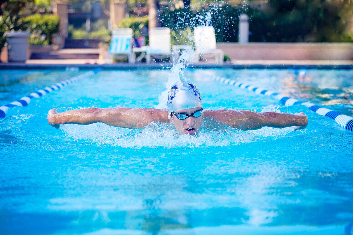 A swimmer wearing goggles and a white cap performs the butterfly stroke in an outdoor pool. Water splashes around their arms, and pool lanes are visible on each side. Sunlight shines on the water, creating a vibrant and dynamic scene.