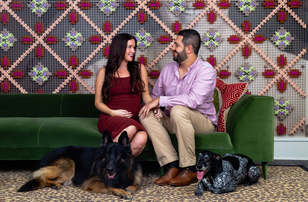 Husband and pregnant wife sit on green couch with two dogs