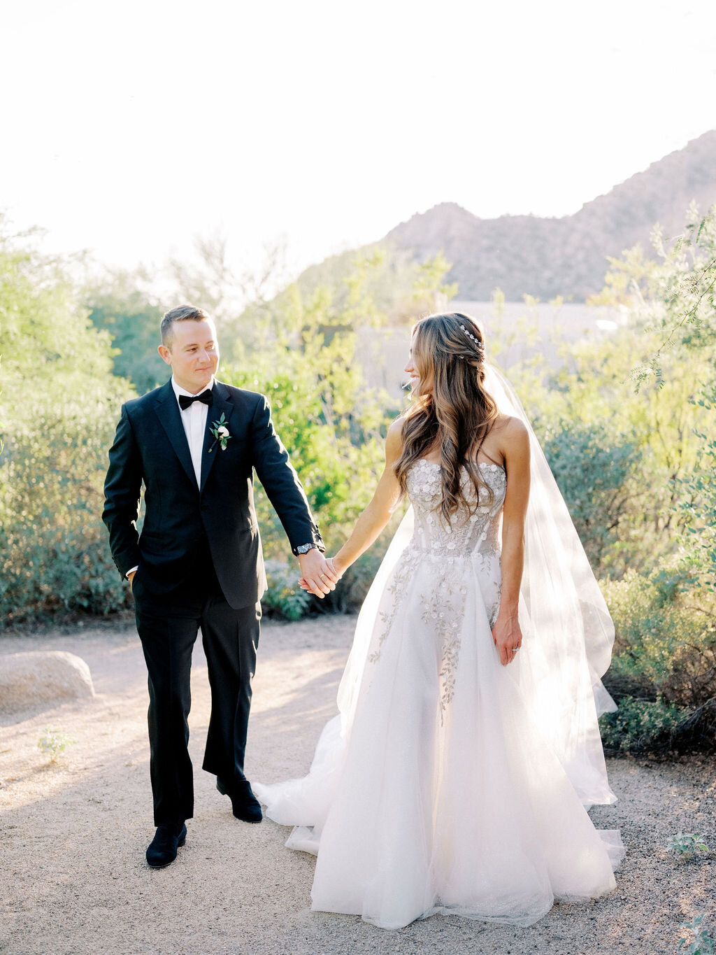 The bride and groom share a loving moment, holding hands under the golden desert sun. A stunning outdoor wedding setting.